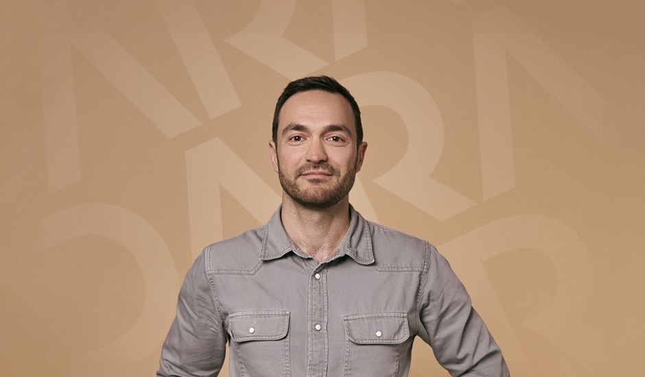 Frontal portrait of Dara Ullrich wearing a grey-green shirt. The beige background features subtle, transparent DARA typography.