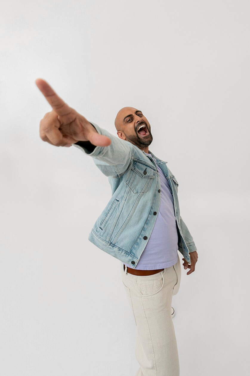 Man smiling sitting on stool wearing denim jacket and cream pants, raising hand with foot extended toward camera against white backdrop.