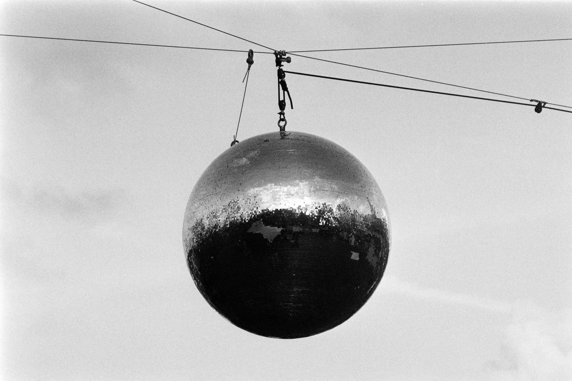 Mirror ball disco sphere suspended from overhead cable against pale grey sky, reflective tiled surface showing distorted landscape fragments