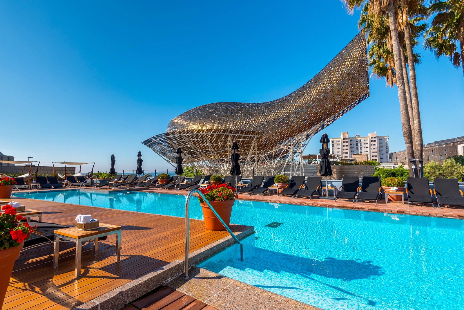 A large, golden, wave-shaped sculpture towers over a turquoise swimming pool lined with lounge chairs and palm trees.