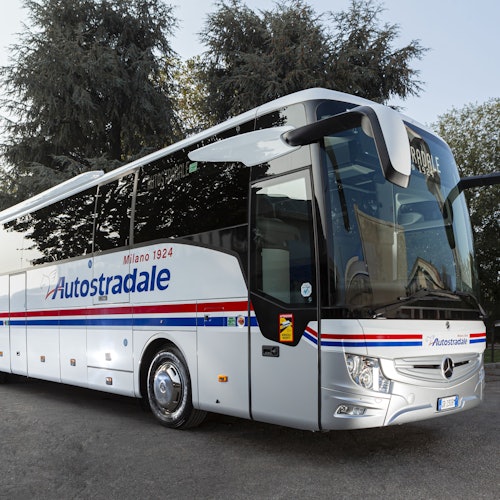 一輛白色豪華長途巴士，上面有「Autostradale」品牌標誌，停在一條後面有高大樹木的道路上。