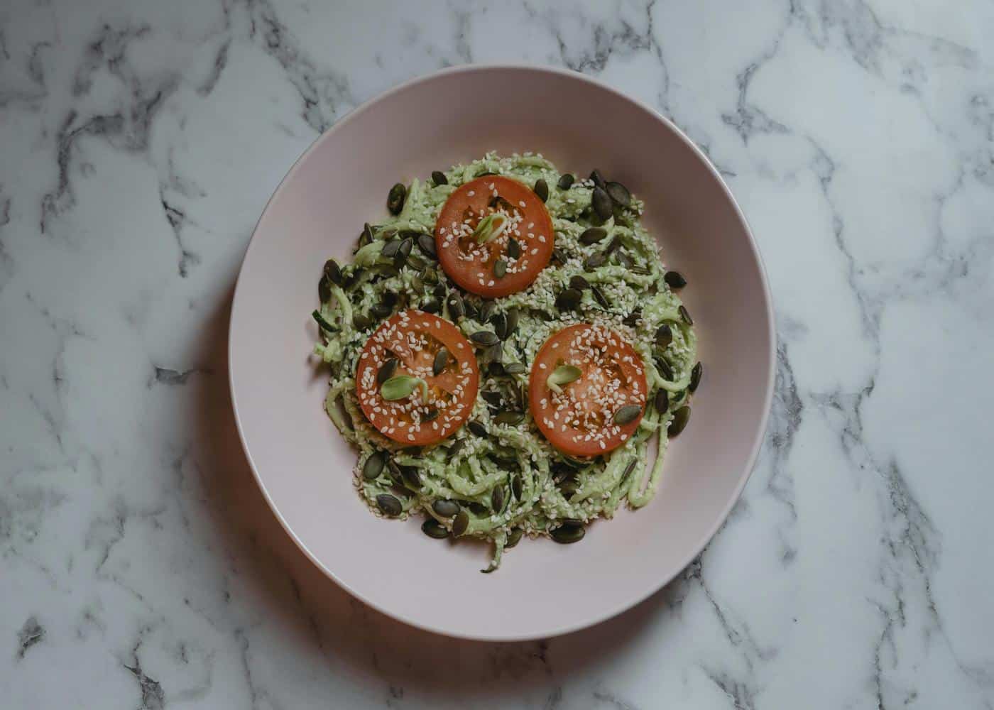 Zucchini noodles on a pink bowl with seeds and tomatoes on top
