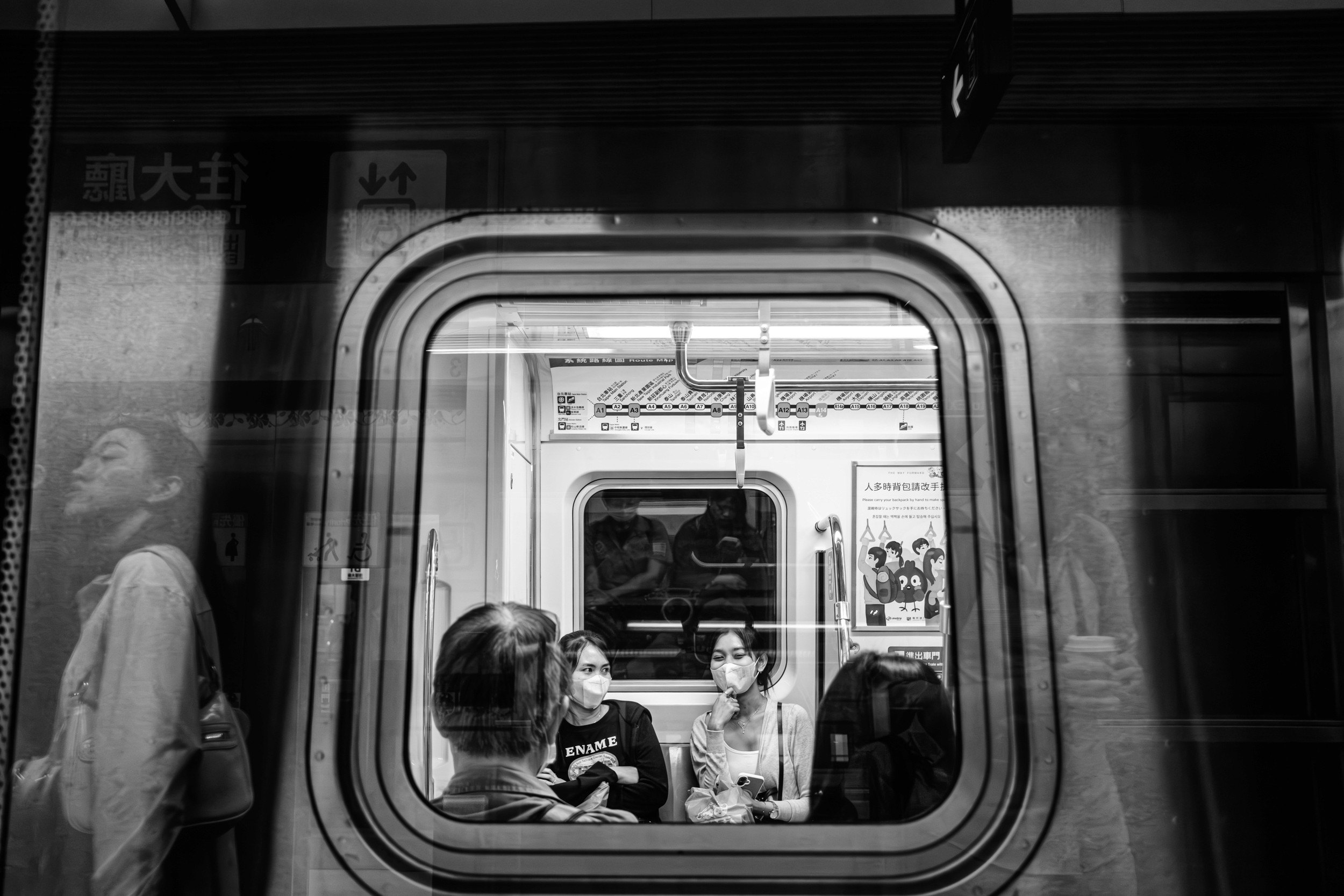 Black and white photograph of commuters visible through a train window layered with reflections from the platform.