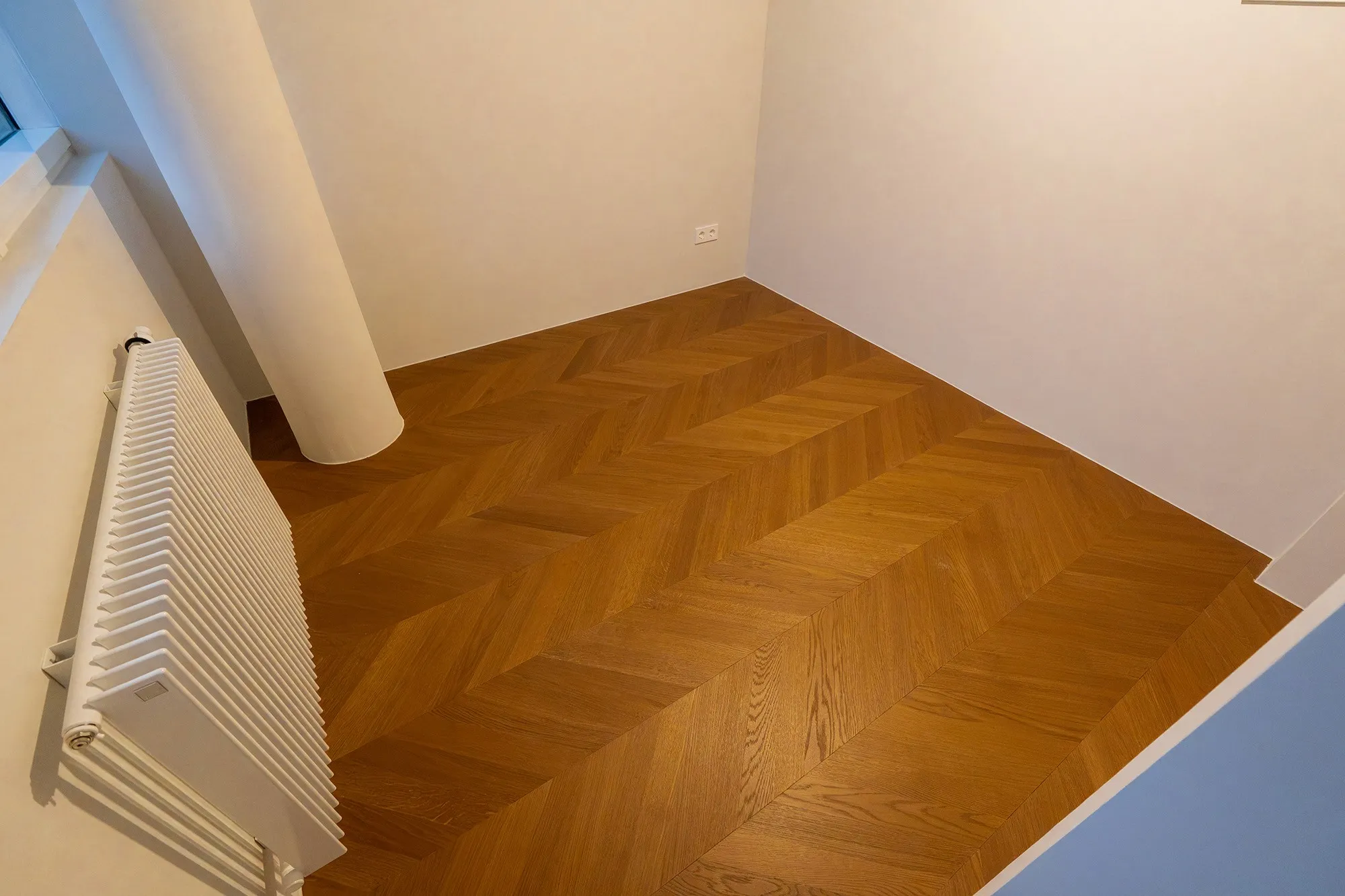 Detailansicht eines präzise verlegten Chevron-Parkettbodens entlang einer Wand und Säule