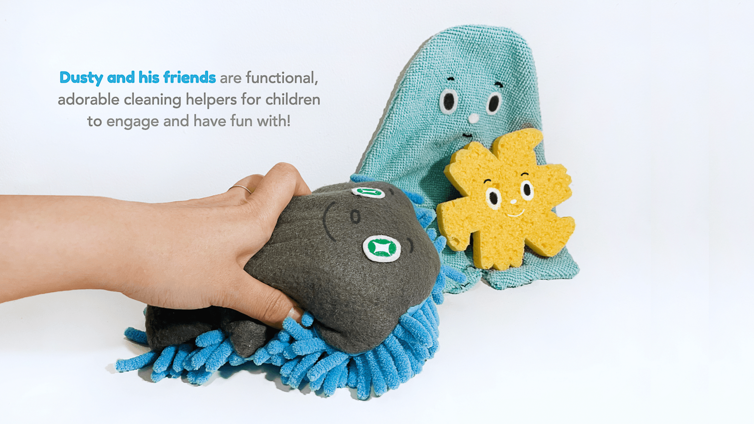 A hand holds a dark gray plush toy with blue fringe and a face with green eyes and a smiling mouth. Beside it, a teal plush toy with eyes and a smiling face is partially visible. A yellow plush star-shaped toy with facial features is positioned near the teal plush. The background is plain white. Text in the top left corner reads “Dusty and his friends are functional, adorable cleaning helpers for children to engage and have fun with!”