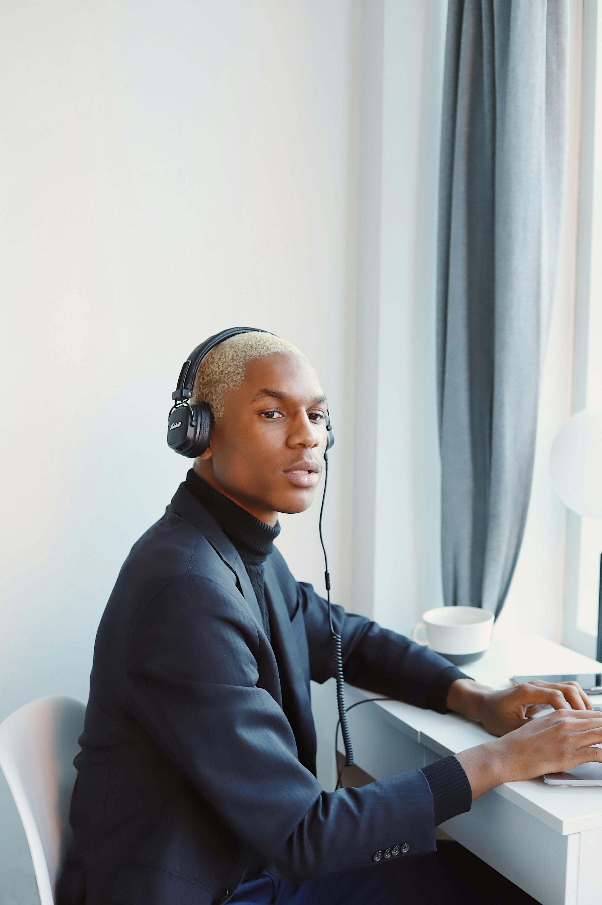 A profile view of the man at his desk wearing the dark blazer and headphones, looking toward the camera with a neutral expression while his hands rest on the laptop keyboard.