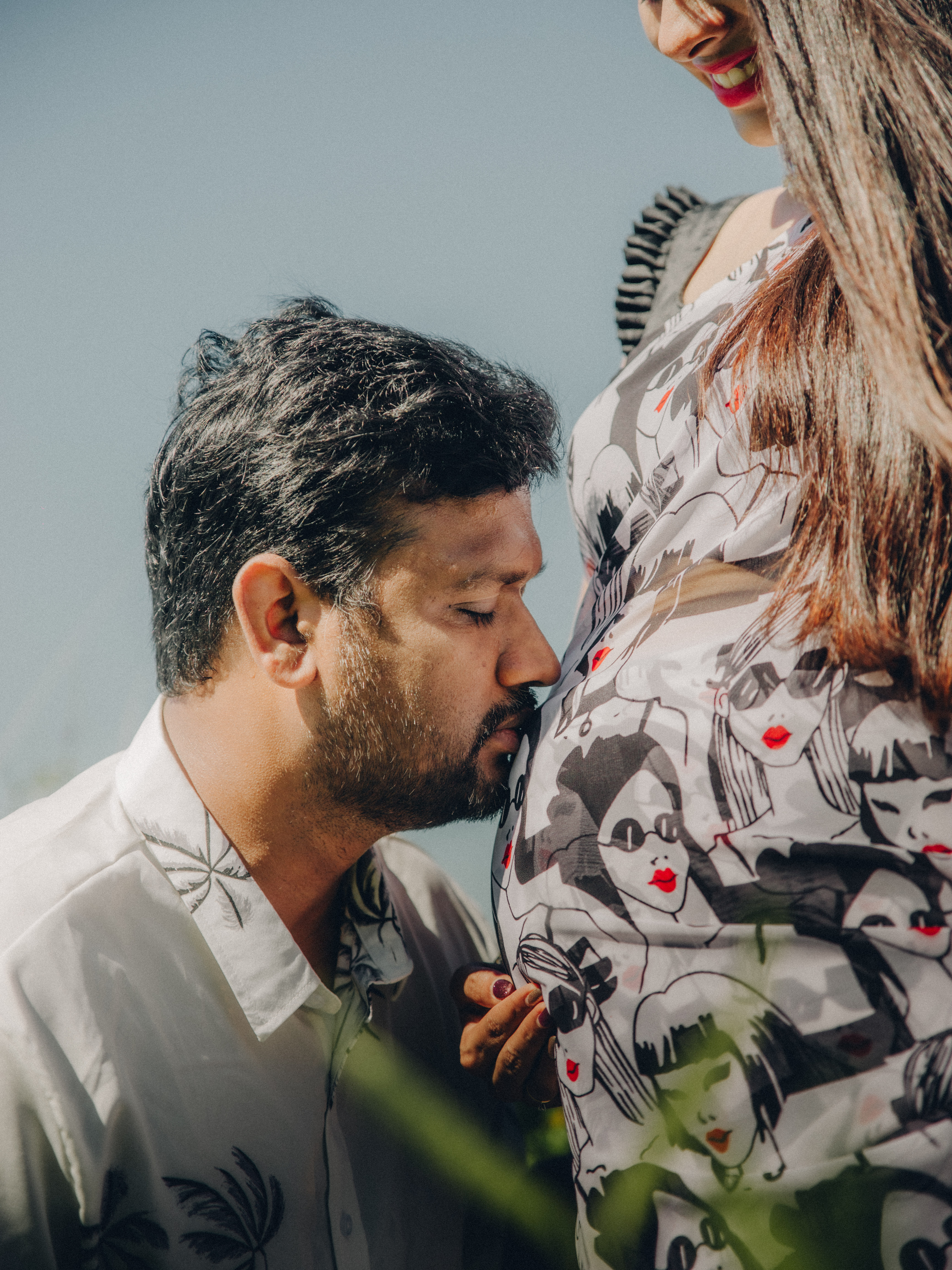 Husband tenderly kissing his pregnant wife's baby bump in an outdoor garden setting.