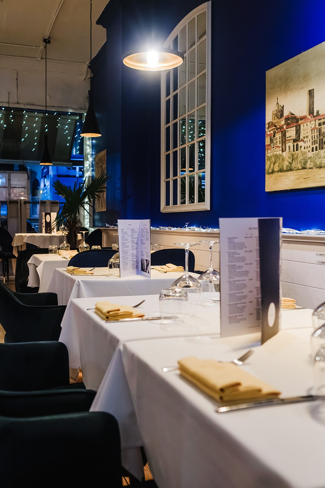 A view across a softly lit restaurant dining room with neatly set tables covered in white cloths, folded napkins, upside-down wine glasses and upright menus. The room features deep blue walls, hanging pendant lights, decorative plants and framed artwork, creating a warm and elegant atmosphere.