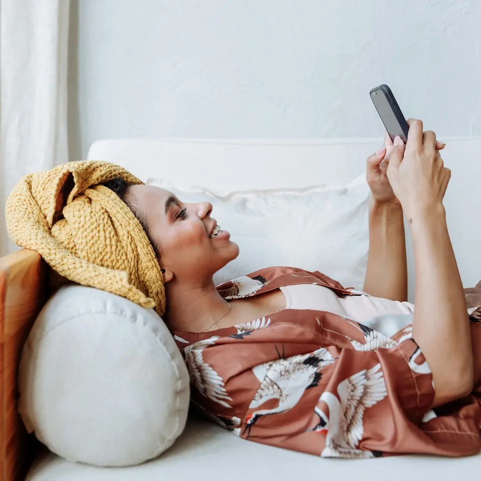 A woman relaxing on a couch at home, smiling while looking at her phone in a calm, unhurried moment.