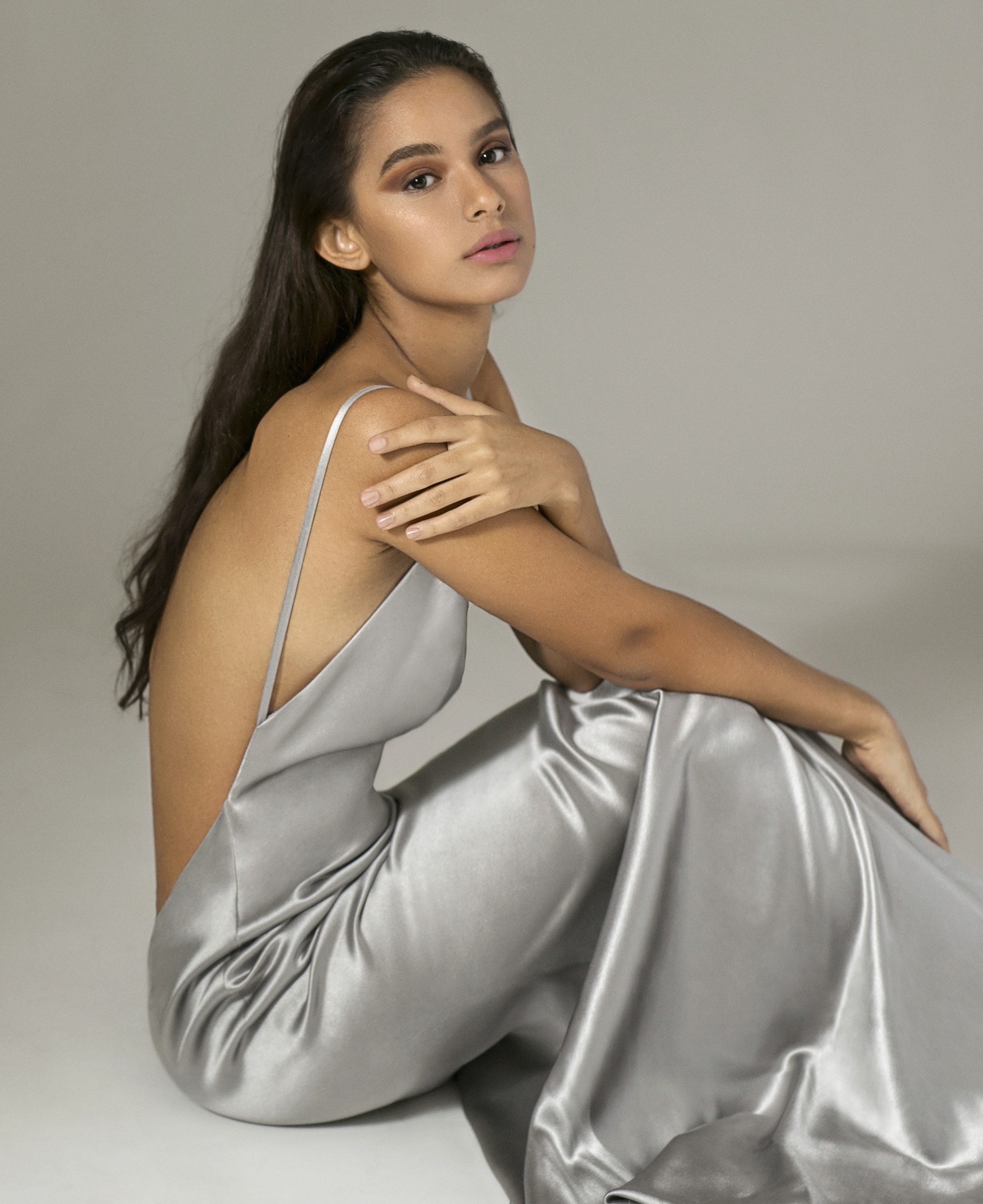 Genesis wearing a silver satin dress by Ecliptica Atelier, seated elegantly with a serene expression, photographed by Carlos Javier.