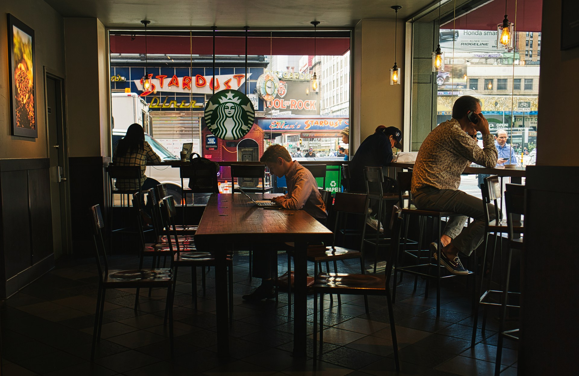 A coffee shop with a warm, cozy atmosphere, featuring several people working and relaxing. One person is seated at a wooden table using a laptop, while others are sitting by the large windows, some on their phones. Outside, bright neon signs and a bustling city street are visible through the glass, including a Starbucks logo and various storefronts.