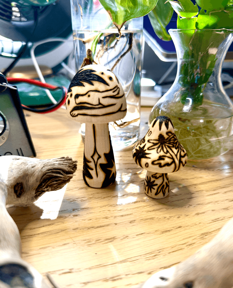 Two wood burned mushrooms. The taller mushroom has a dog chasing a shooting star with starbursts patterning the rest of the mushroom. The smaller mushroom is covered in Black Eyed Susans and star bursts. The mushrooms are surrounded by plants, drift wood, and the colwood pro 2 wood burner.