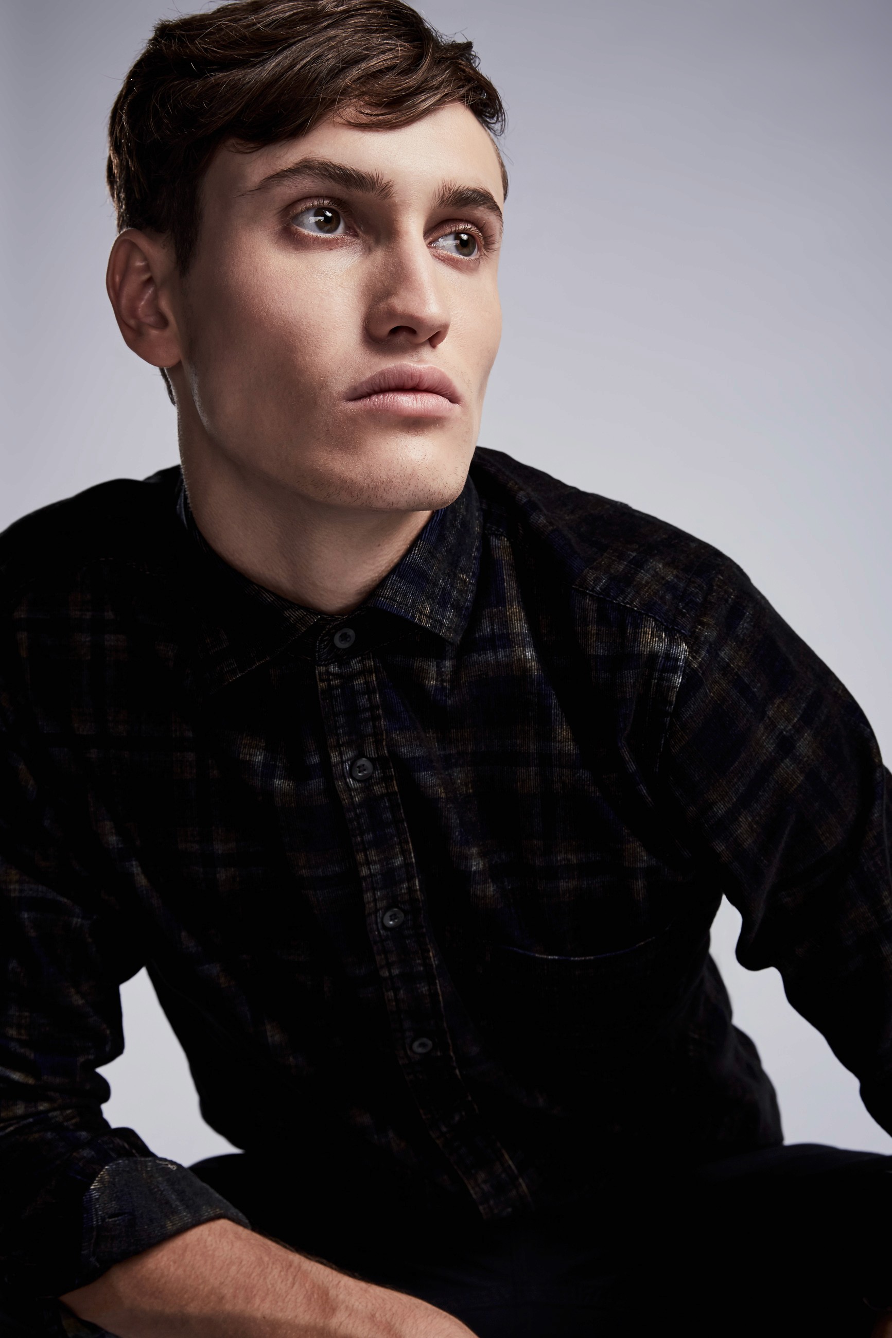 A close-up of a male fashion model wearing a dark patterned shirt sitting and leaning forward while looking off to the side.
