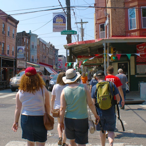 A group of people walking on a sunny street lined with brick buildings, colorful flags, and a street sign reading "Christian St."