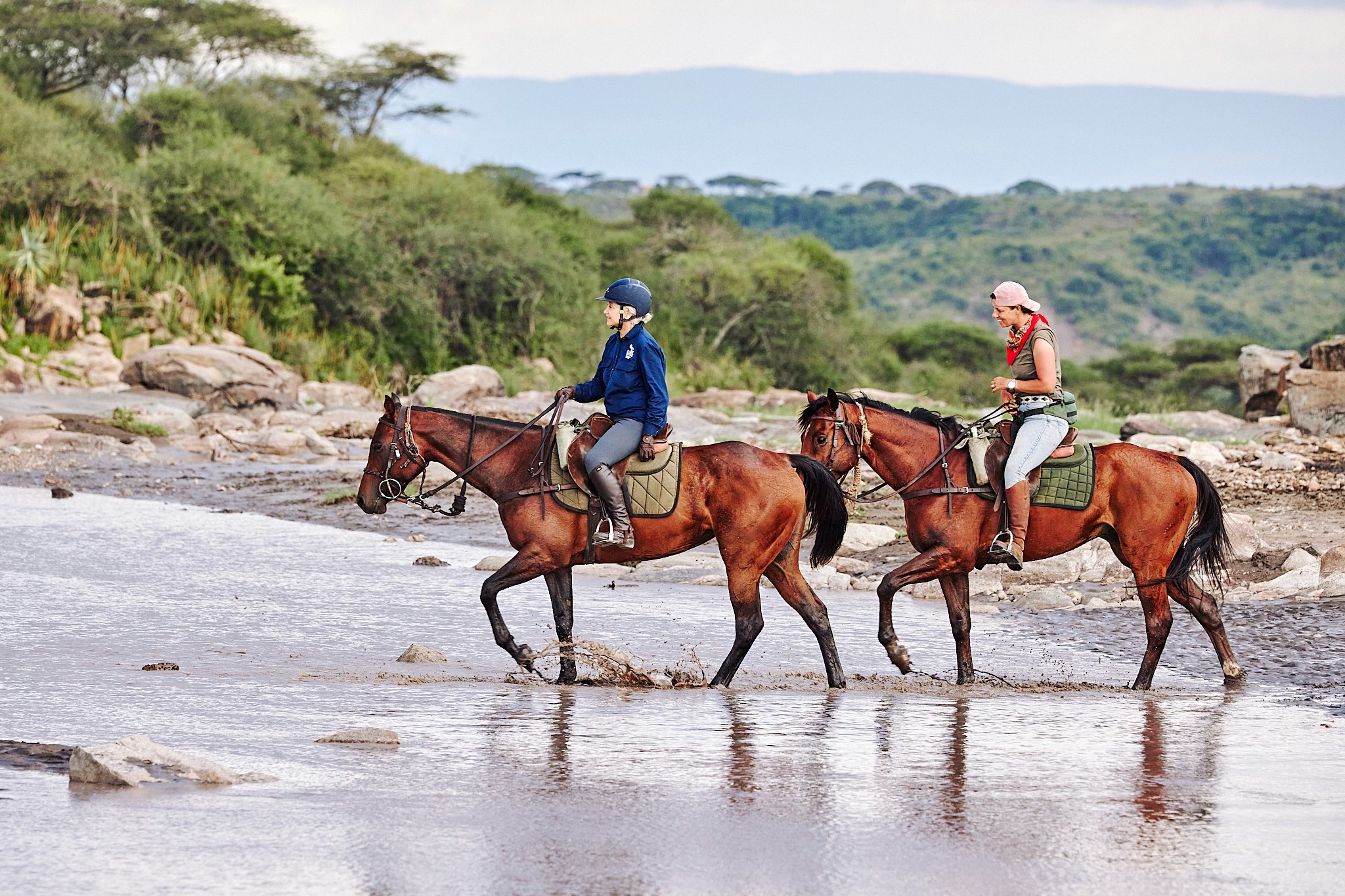 Två ryttare på bruna hästar korsar en flod i Serengeti, omgivna av grönskande landskap och kullar i bakgrunden.