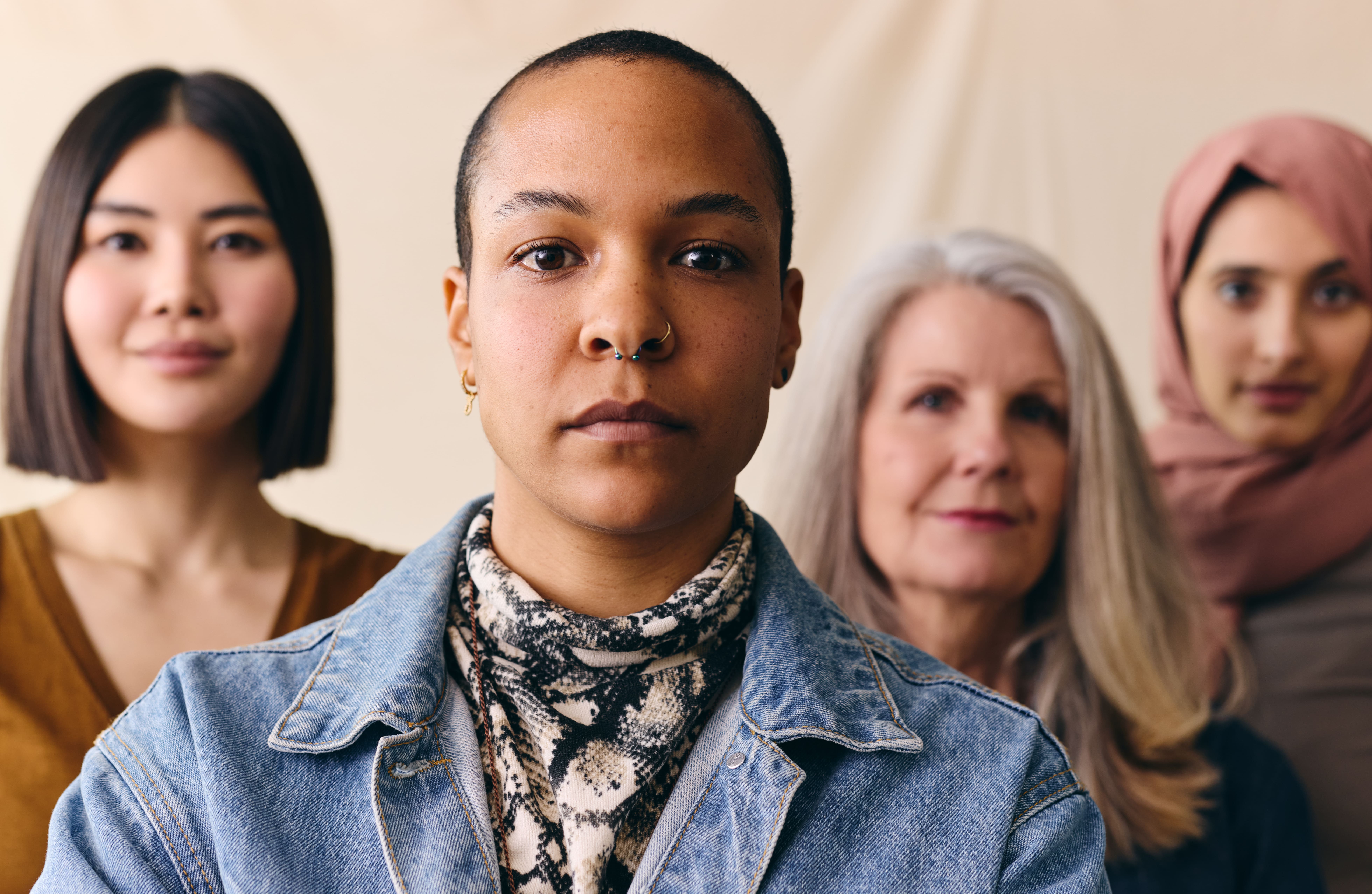 A imagem mostra quatro mulheres de diferentes origens e idades, alinhadas lado a lado, olhando diretamente para a câmera. A mulher à frente, no centro, tem cabelos raspados, usa um piercing no nariz e veste uma jaqueta jeans com uma blusa de estampa de cobra. Sua expressão é séria e confiante. Atrás dela, as outras três mulheres têm expressões neutras ou levemente sorridentes. À esquerda, uma mulher com cabelo escuro e liso está usando uma blusa marrom. No lado direito, uma mulher mais velha com cabelo grisalho veste uma blusa preta, e à extrema direita, uma mulher com um lenço cor de rosa está parcialmente desfocada. A imagem transmite diversidade e força, destacando a individualidade de cada uma das mulheres, mas ao mesmo tempo sugerindo unidade entre elas.