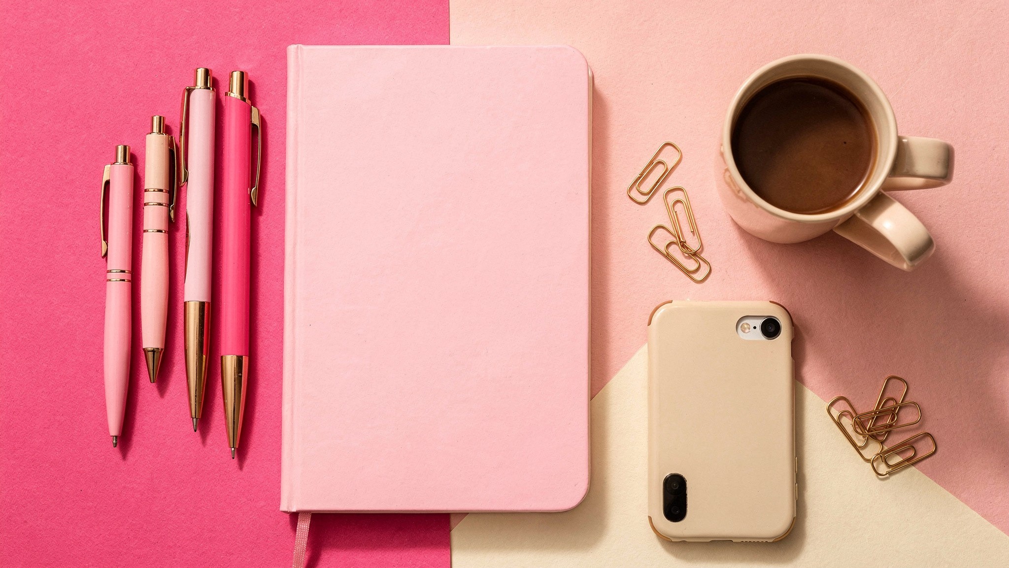 Pink flat lay of a creator's desk with notebook, pens and phone for planning Instagram hashtags