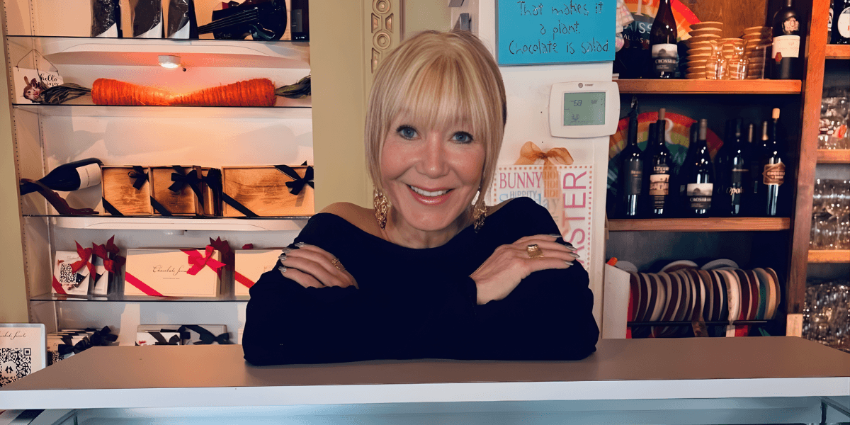 The image shows Suzanne Palmlund, the 88-year-old founder of Enjoy Fun Fudge, smiling behind her store counter, showcasing her business.