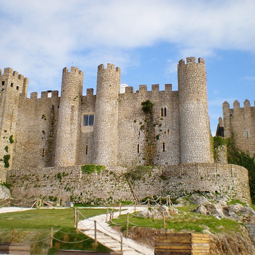 قلعة حجرية بأبراج دائرية وجدران متعرجة ونوافذ صغيرة، محاطة بمنطقة عشبية وحبال. سماء زرقاء فوقها.