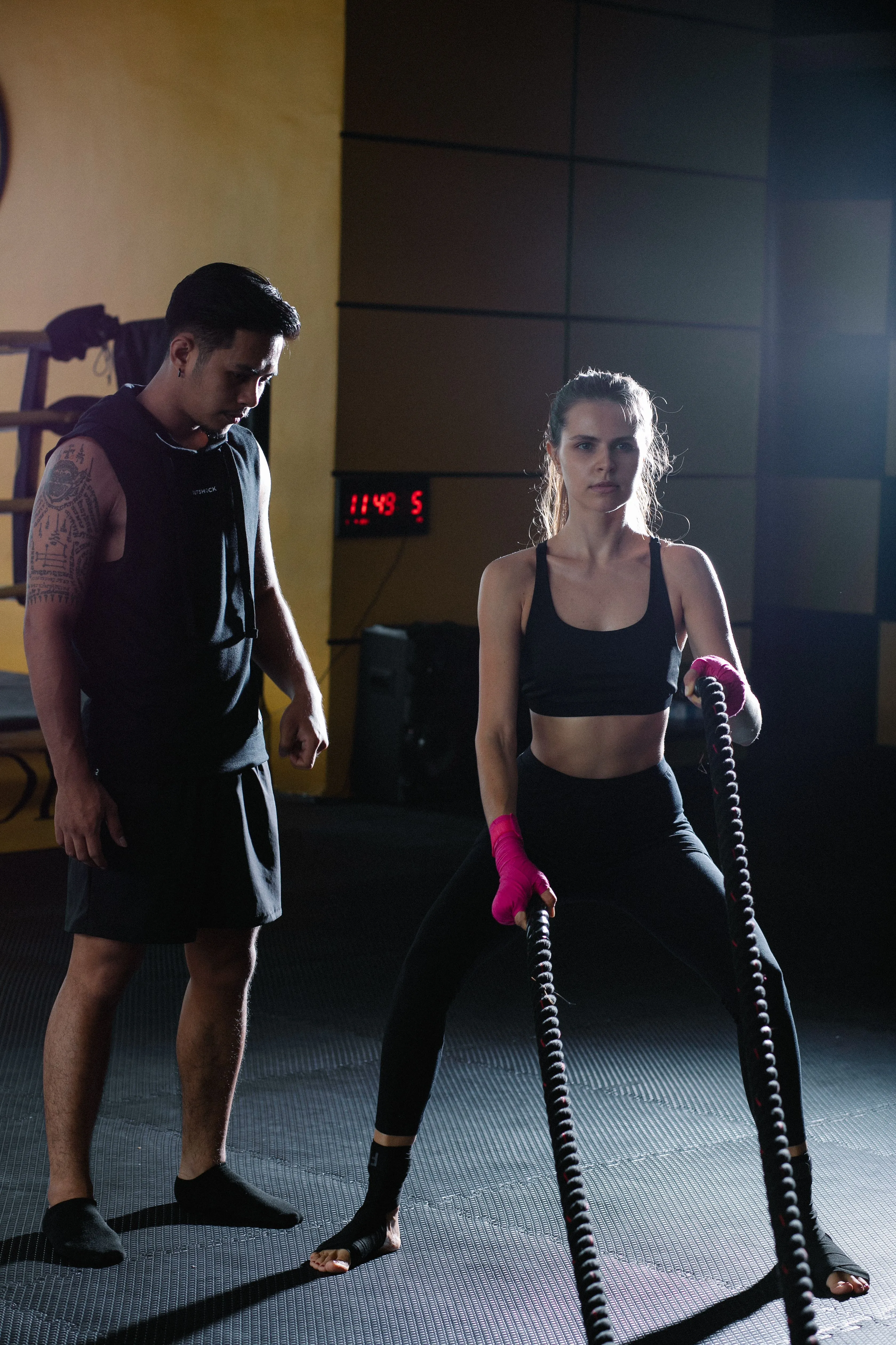 A woman in athletic wear confidently performs a battle ropes exercise in a dimly lit gym, with a trainer observing her form.