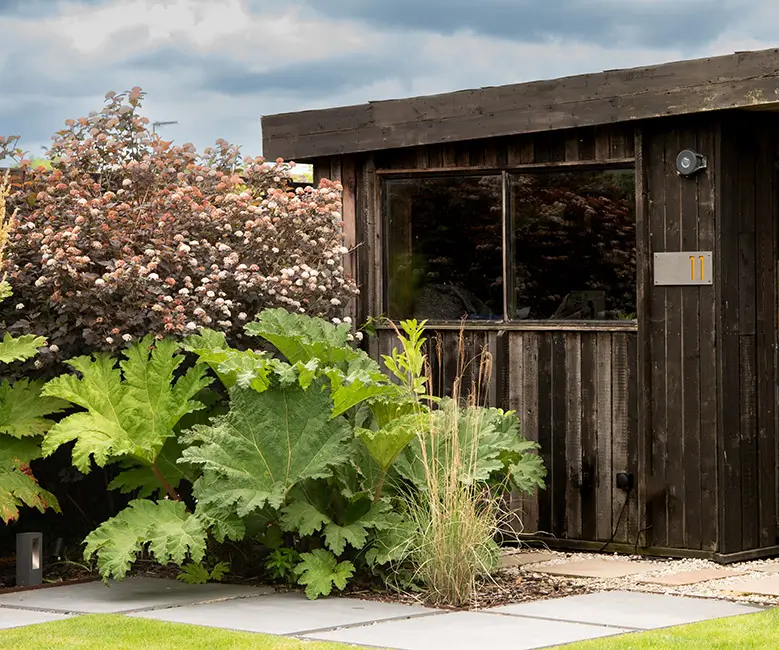 A wooden shed surrounded by lush greenery and colorful plants under a cloudy sky.