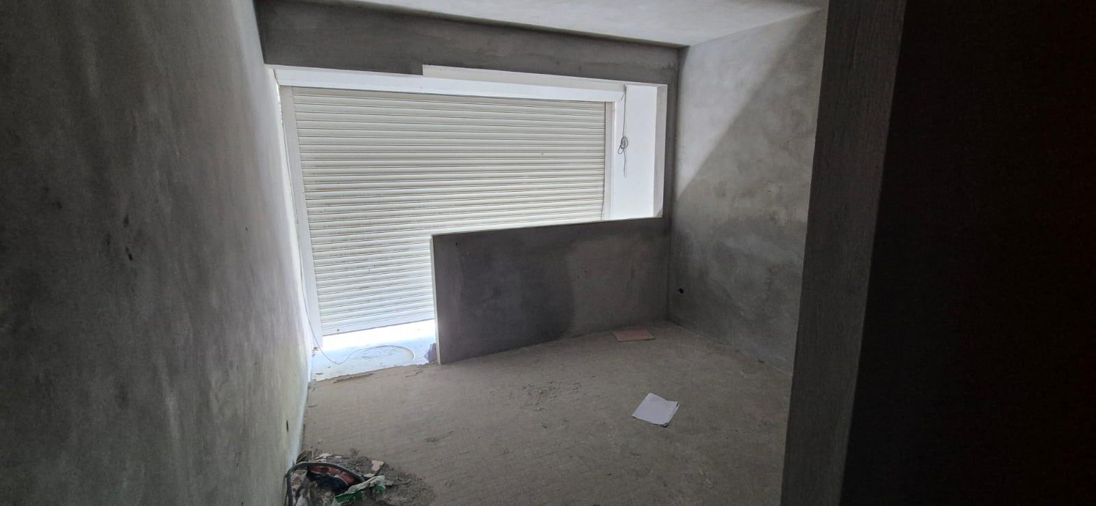 Pièce faiblement éclairée en cours de construction, avec des murs et un sol gris inachevés, et une fenêtre à volet blanc partiellement ouverte. Câbles et papiers éparpillés. Ambiance minimaliste, travaux en cours.