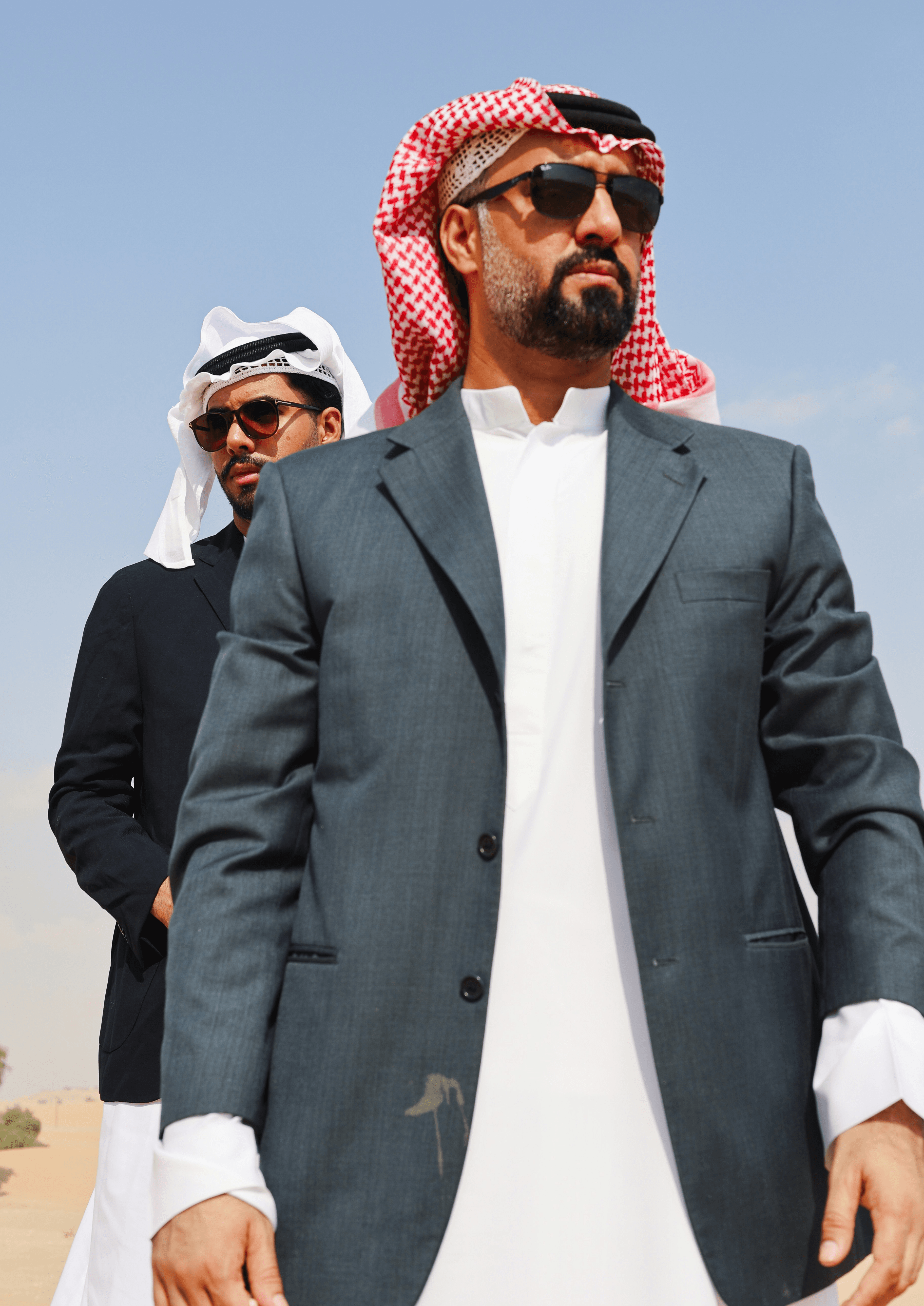 A man in a blue bomber jacket stands against a dark background. The lighting is dramatic, highlighting his focused expression. His pose is confident.