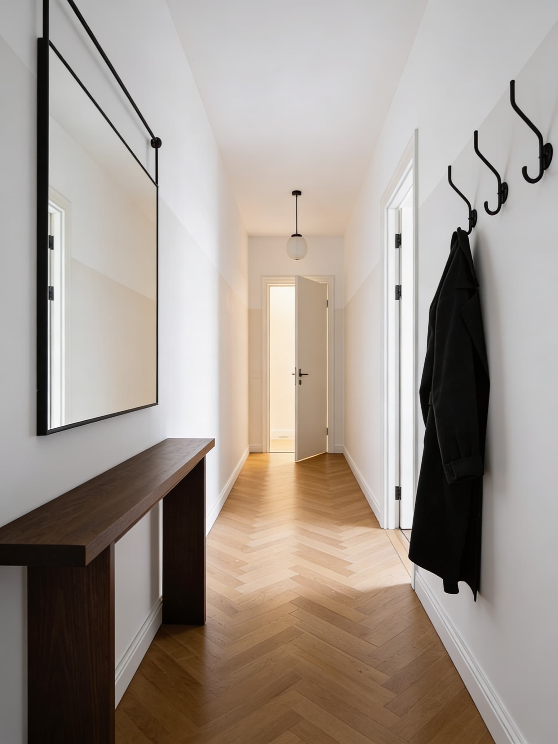 A minimalist hallway interior features white walls, a large mirror with a black frame above a narrow wooden console table, and a black coat hanging from wall hooks, leading to an open door at the end with wooden herringbone flooring.