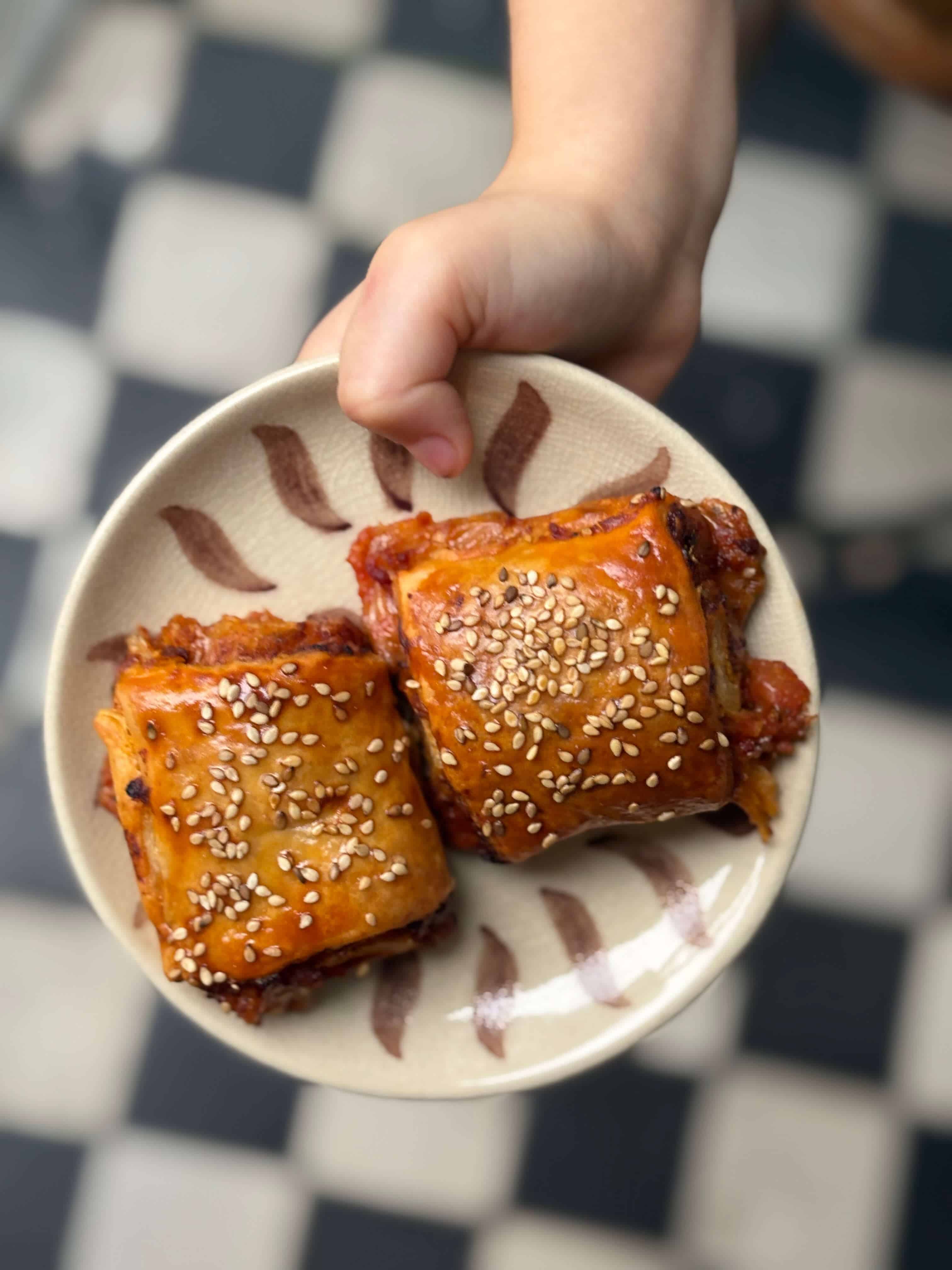 Two homemade pizza scrolls topped with sesame seeds, served on a small ceramic plate held in a child’s hand.