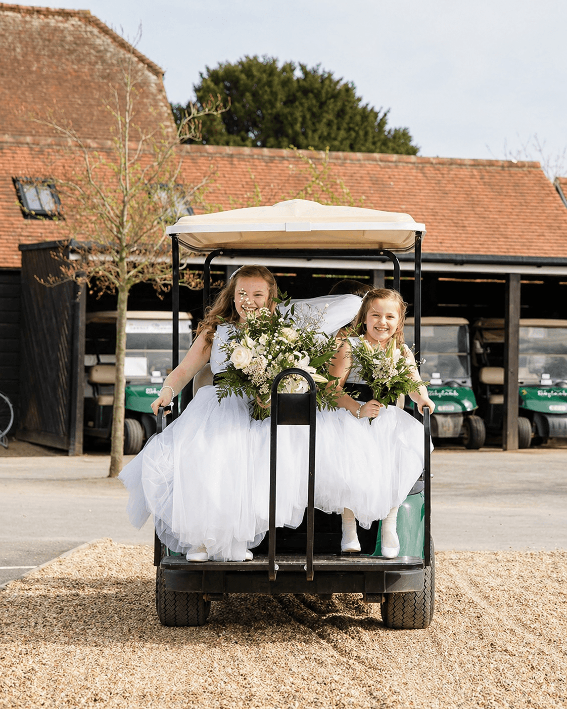 A smiling bride and groom stand holding hands on a green lawn with trees and tall grass in the background at