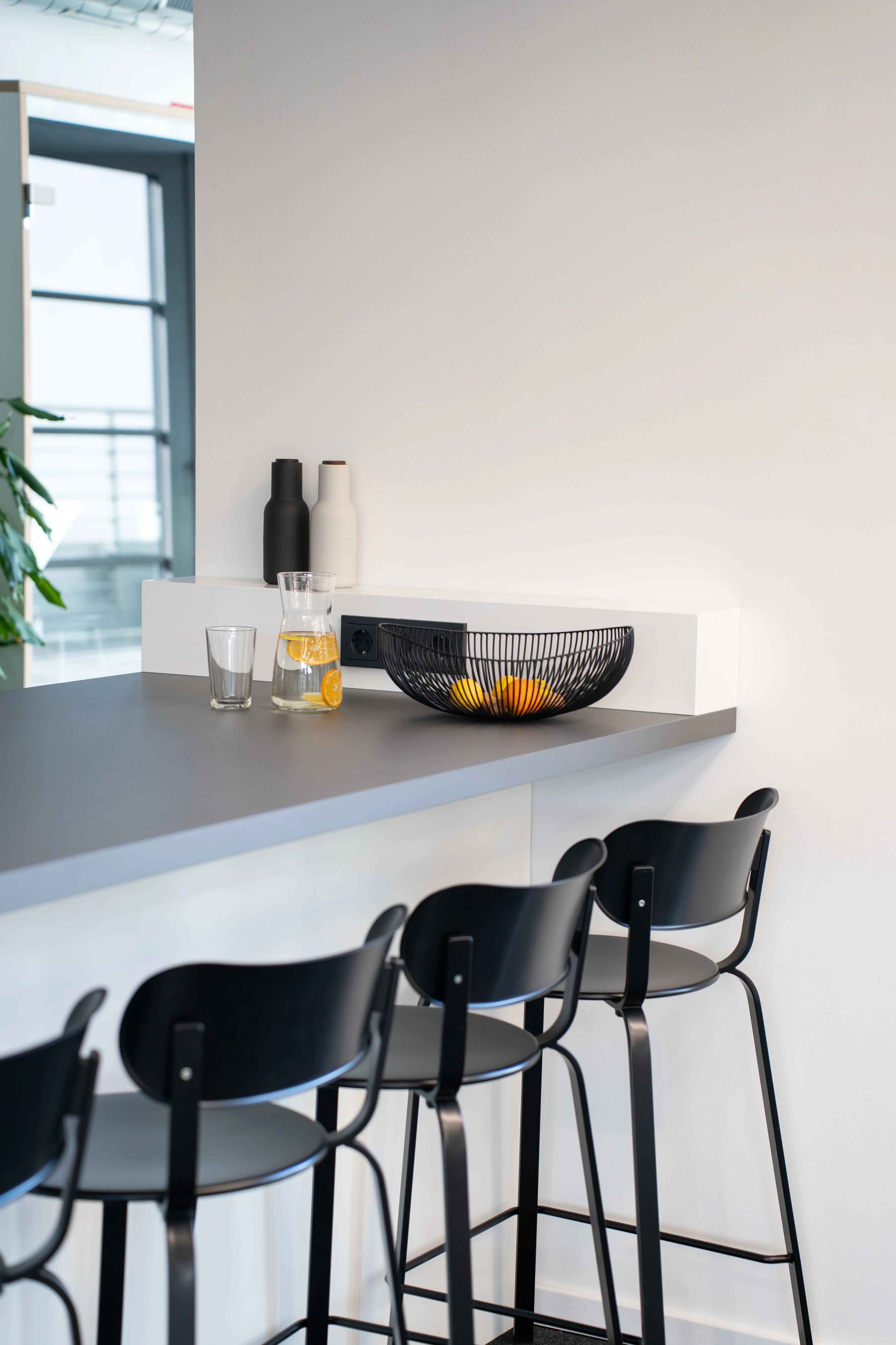 Kitchen counter detail at AI START-UP MUNICH with a grey worktop, black wooden bar stools, a black wire fruit basket, water carafe and ceramic bottles.