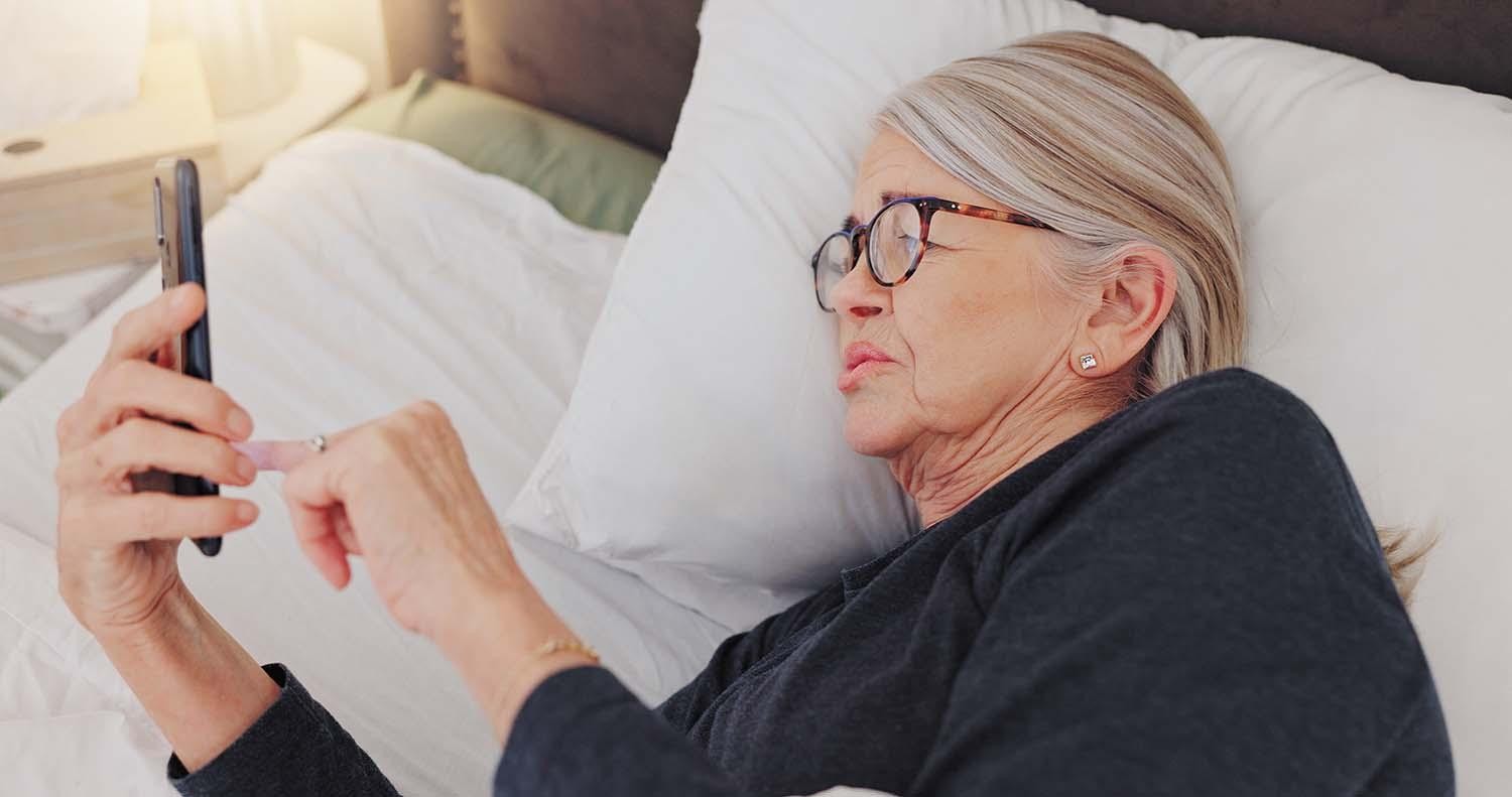 An older woman lying in bed, wearing glasses, using a smartphone with a serious and slightly sad expression, reflecting deep thought or concern.