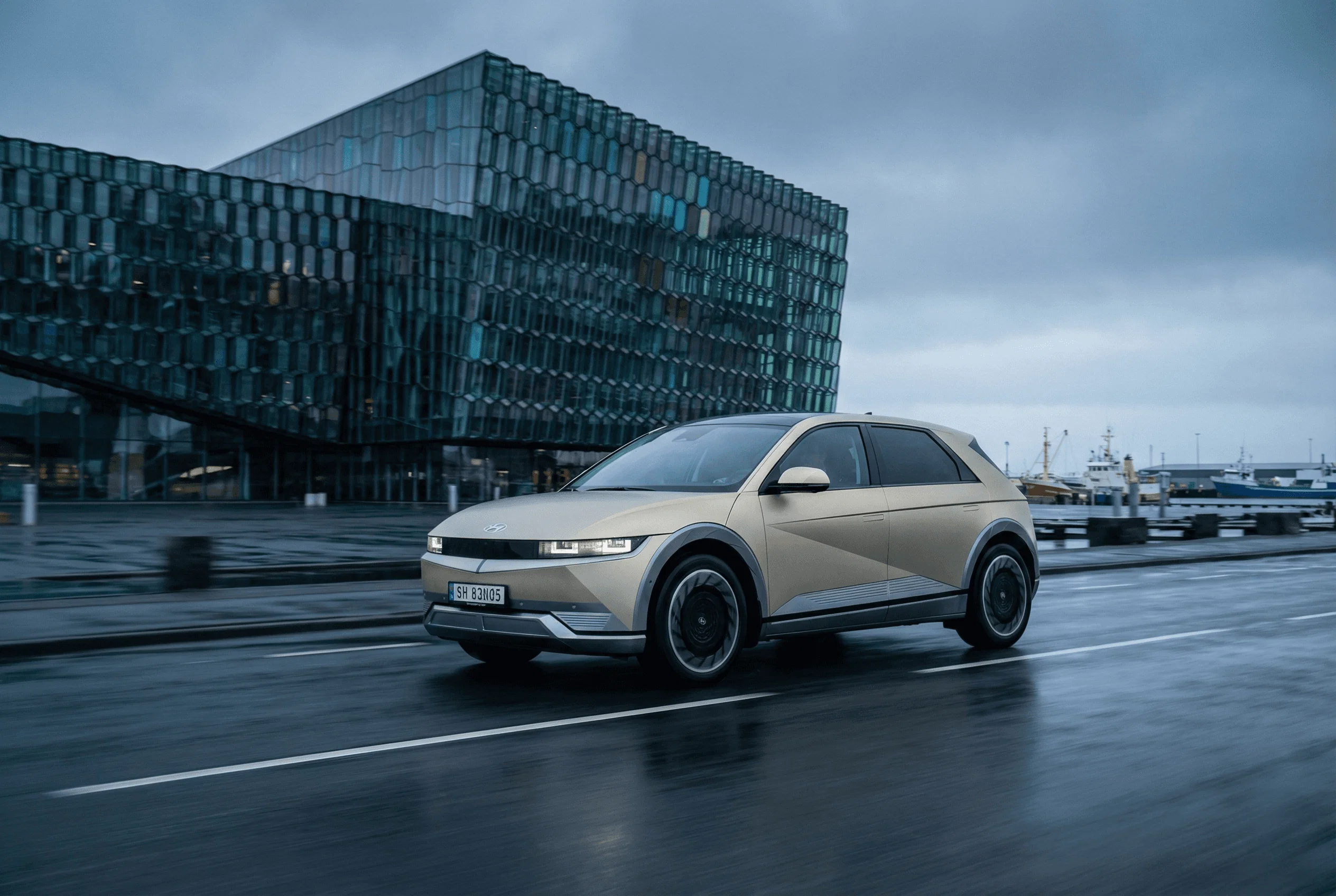 A beige electric SUV parked on wet pavement in front of the glass facade of the Harpa Concert Hall.