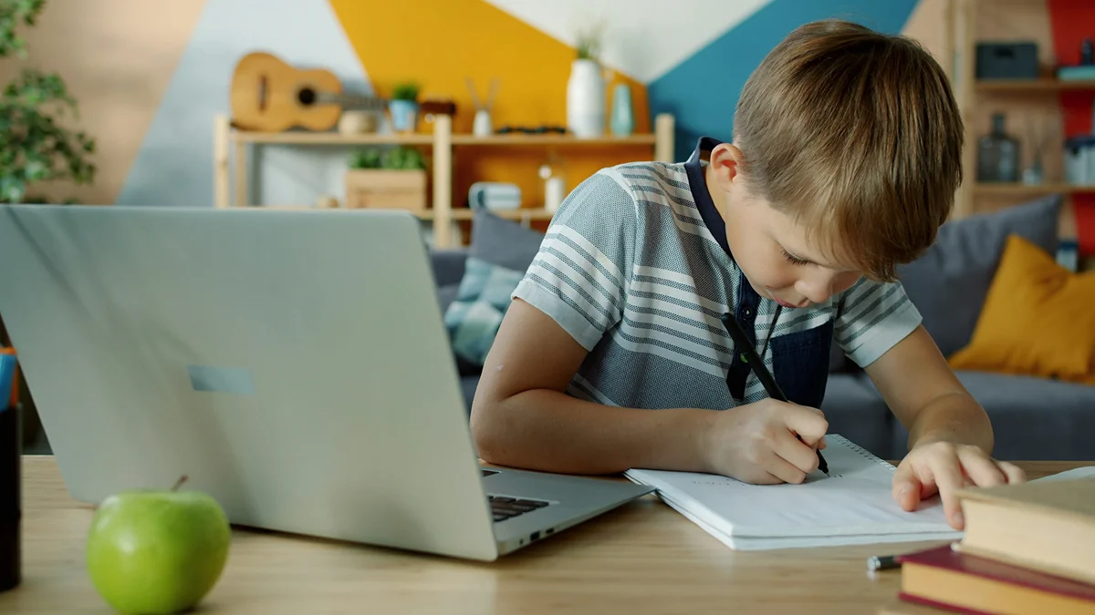 A boy writing in a notebook beside a laptop.