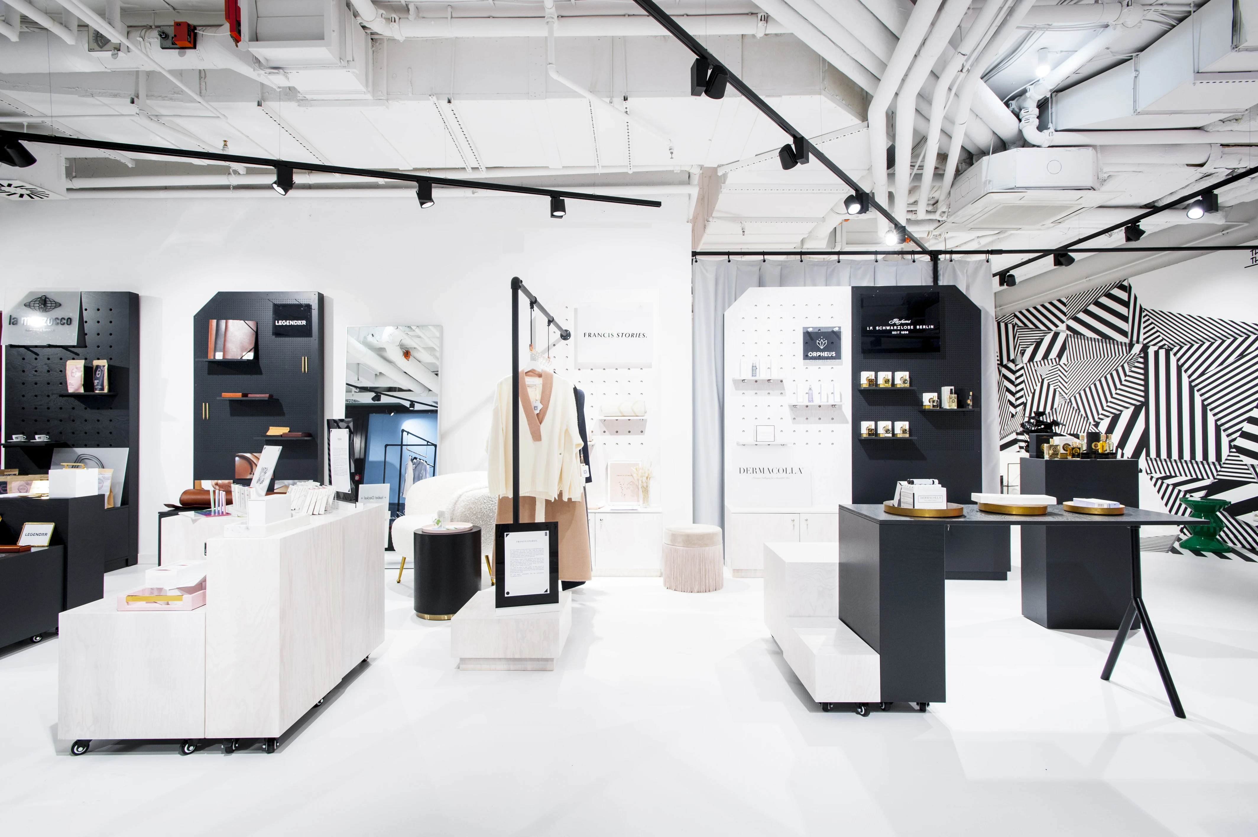 Brand corners at FREIRAUM SHOP with black pegboard displays, white plinths on castors, a clothes rail with knitwear, gold-rimmed poufs and a black-and-white graphic wall.