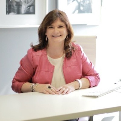 Founder, Angie Marie Grainger, sitting across a desk in an executive office filled with light, smiling with great joy directly at the viewer