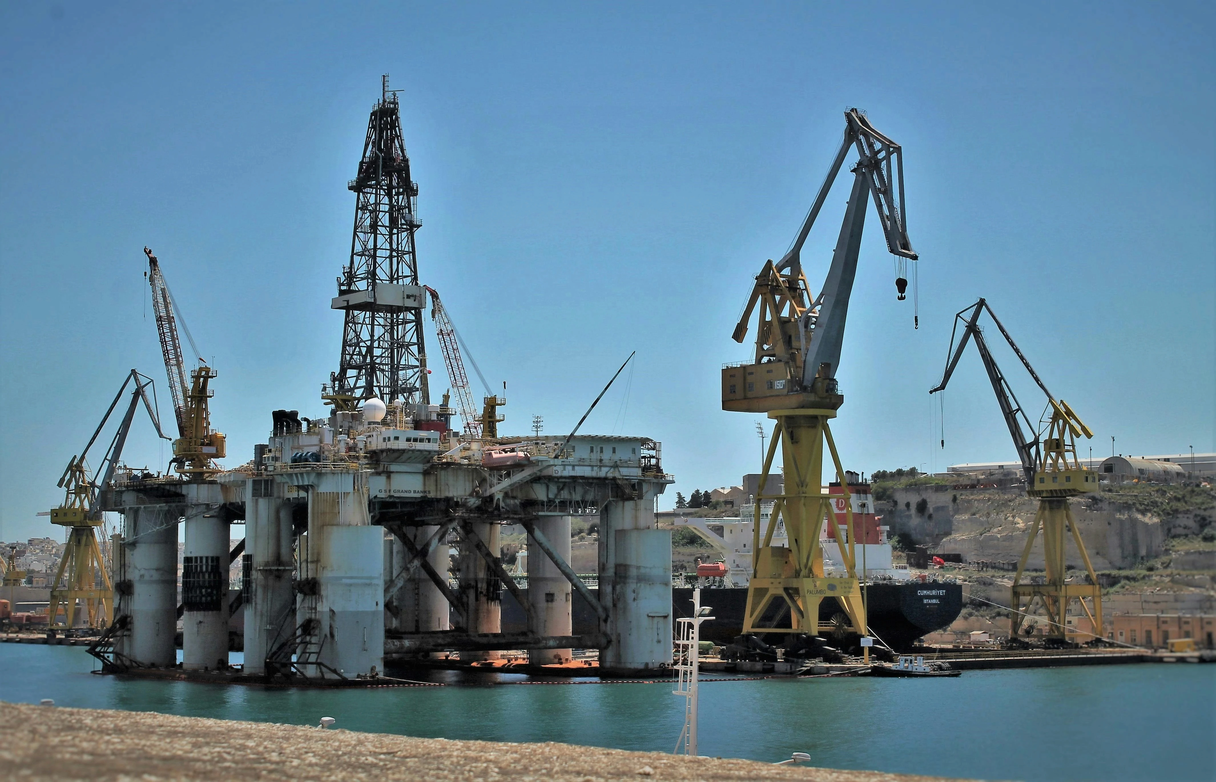 Semi-submersible drilling rig and large yellow shipyard cranes docked in a harbor for maintenance.