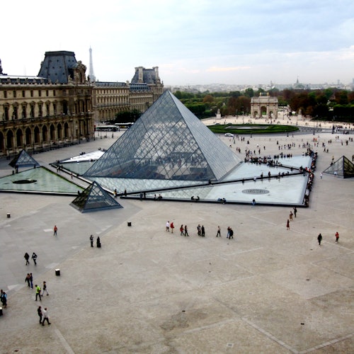 Personas caminando cerca de la entrada de la pirámide de cristal del Museo del Louvre, con los edificios históricos circundantes y el paisaje urbano distante.