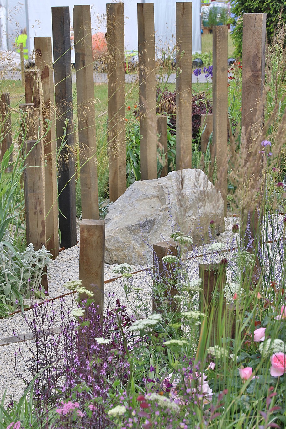 A small stone sits among flowers and plants, surrounded by rustic wooden posts in a garden setting.