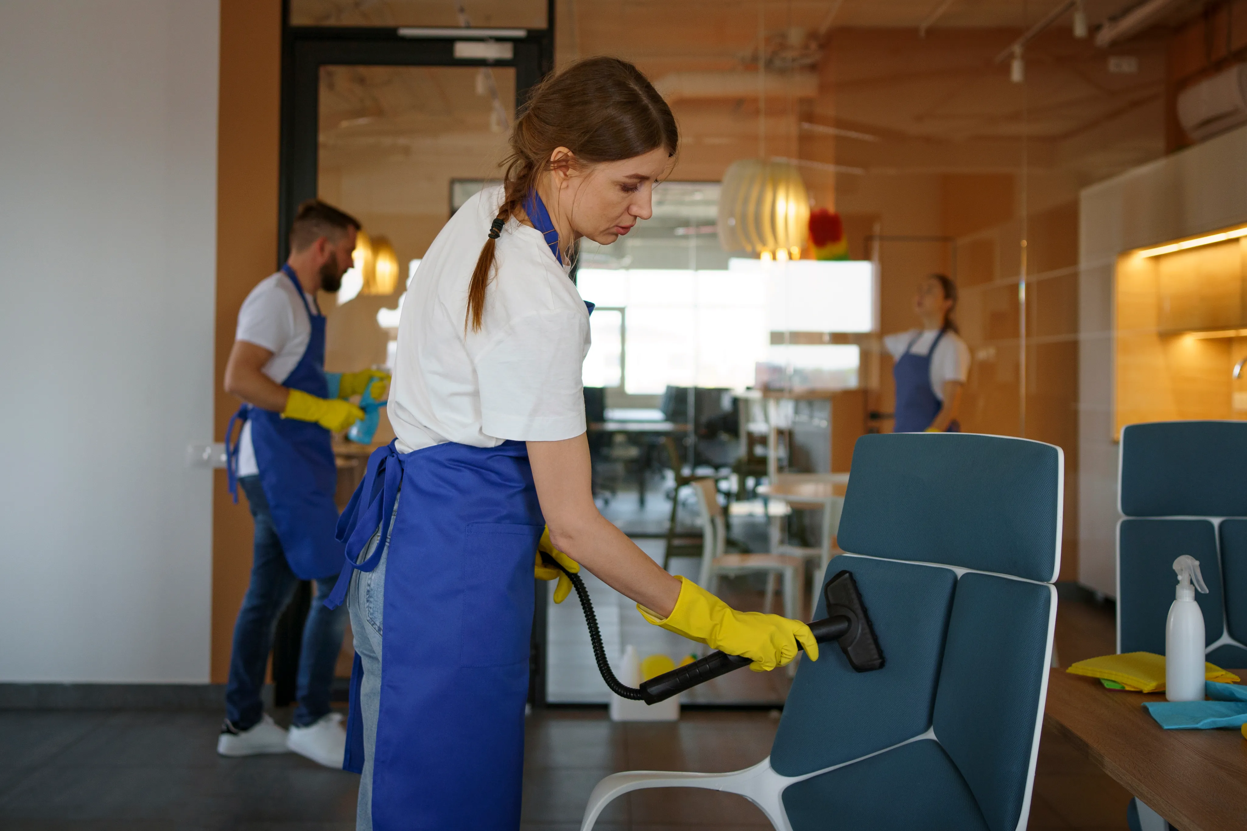Two professional cleaning staff members in blue aprons and yellow gloves deep cleaning office chairs in a contemporary coworking space.