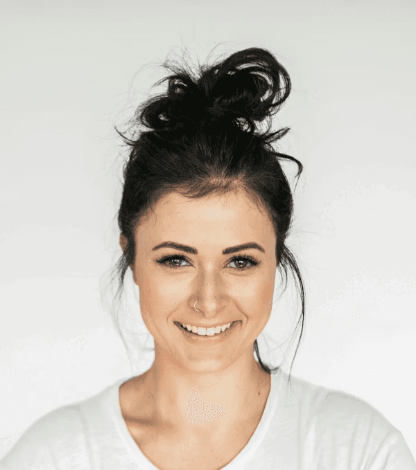 Smiling woman with dark hair in a messy bun and a nose ring, wearing a white t-shirt against a light background.