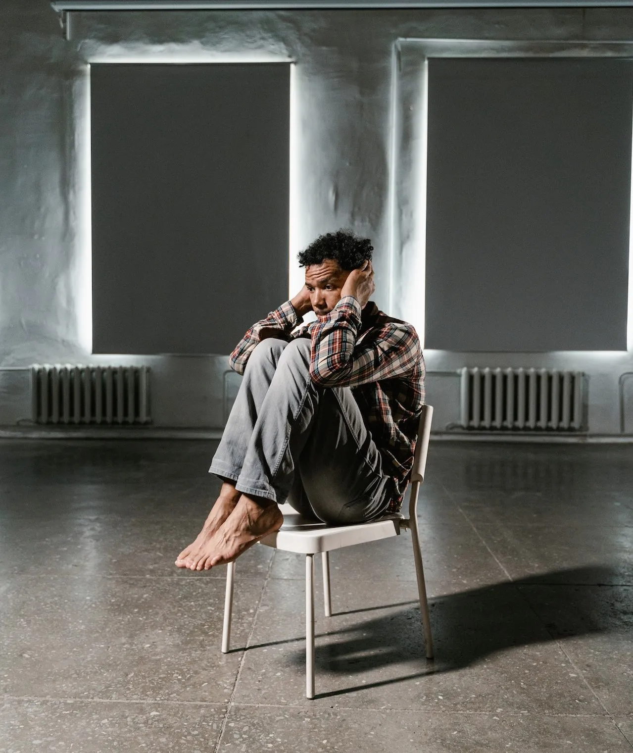 A person sits on a chair in a dimly lit room with large windows, looking contemplative and lost in thought.
(Photo by MART PRODUCTION: Pexels)
