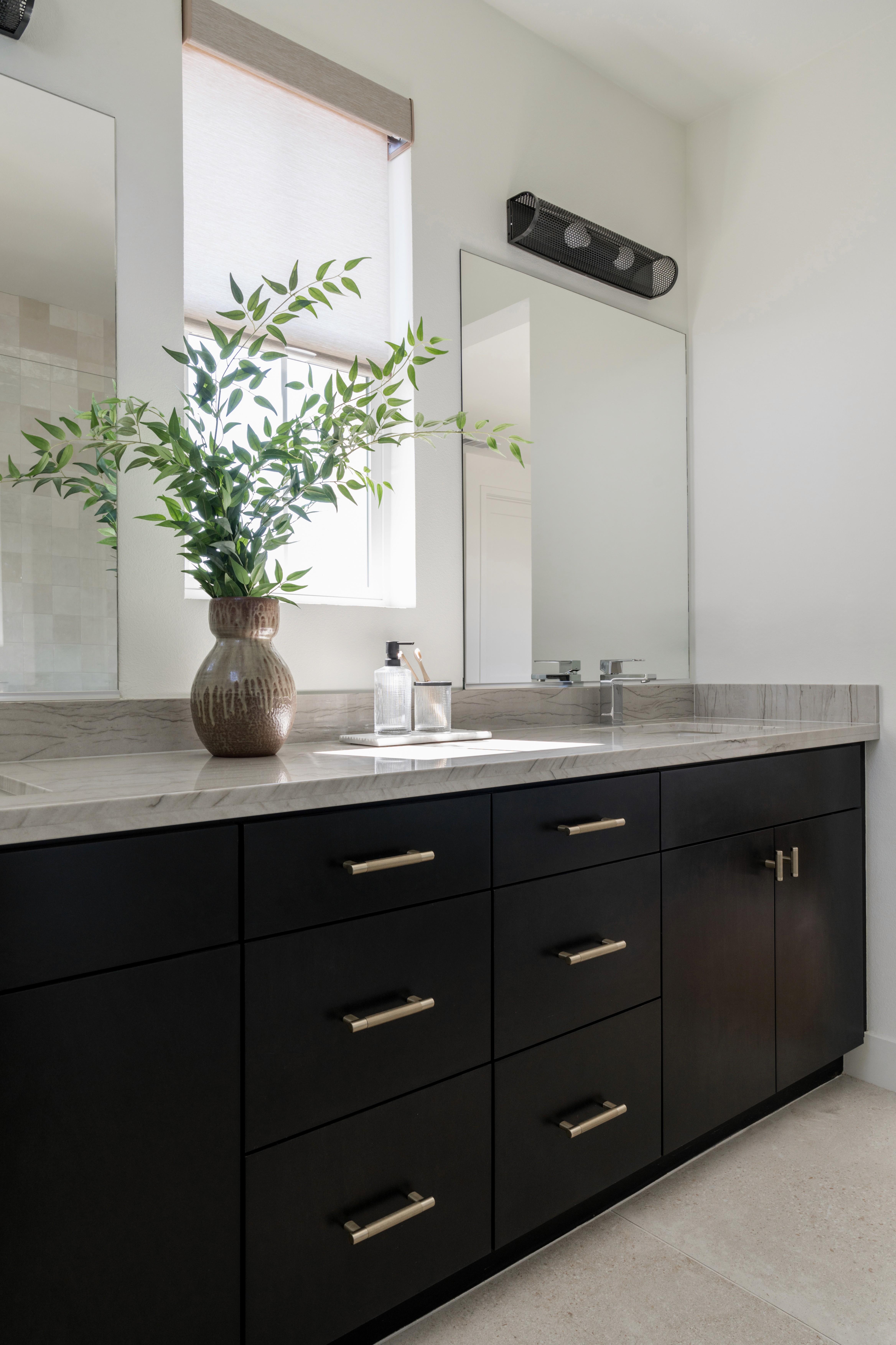 View of one sink in a dual sink vanity bathroom with black cabinetry, gold finishes, roman shades, grey marbled countertop, and chrome faucet