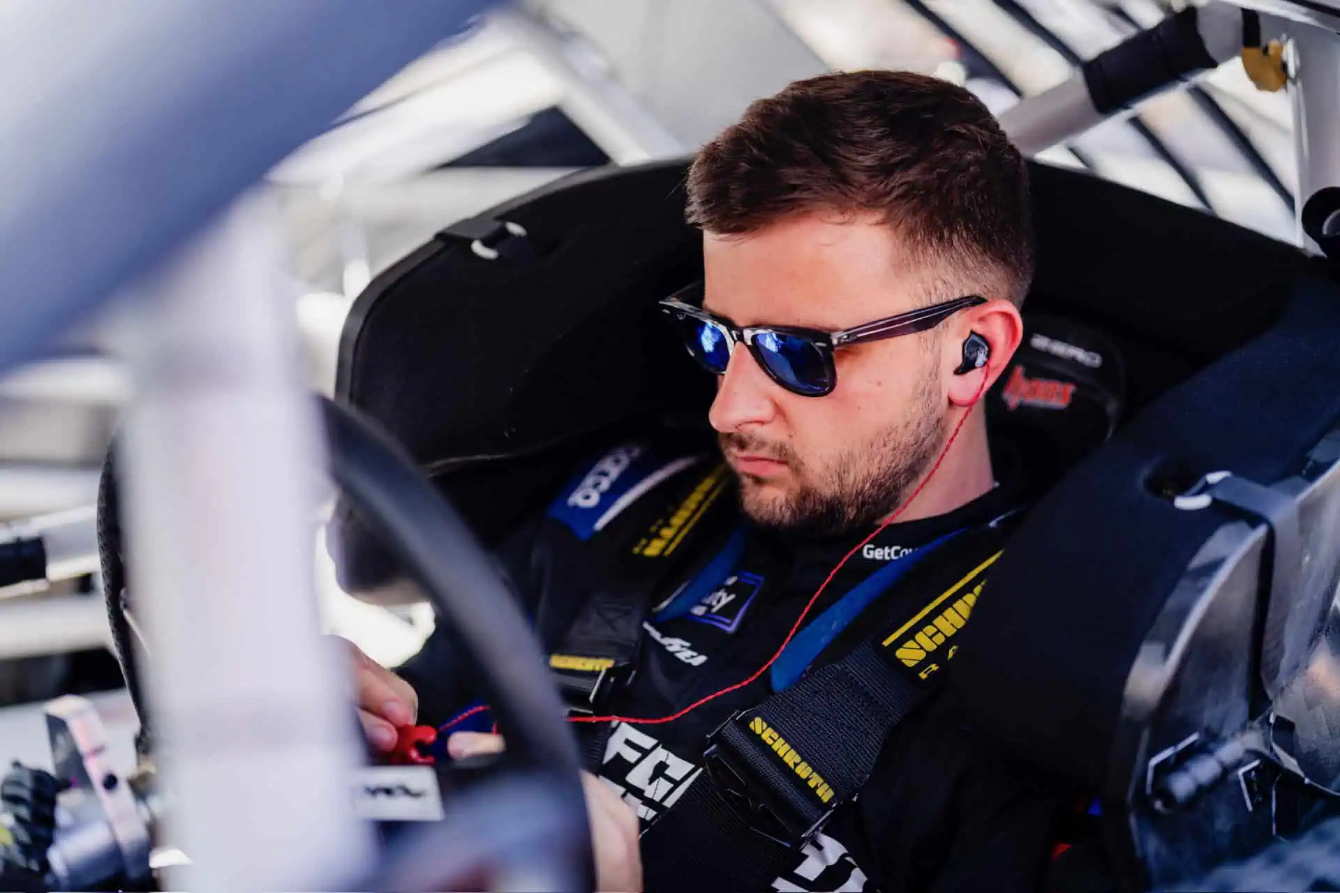 Joe Graf Jr. sitting inside his NASCAR race car wearing sunglasses and headphones, preparing before a race.