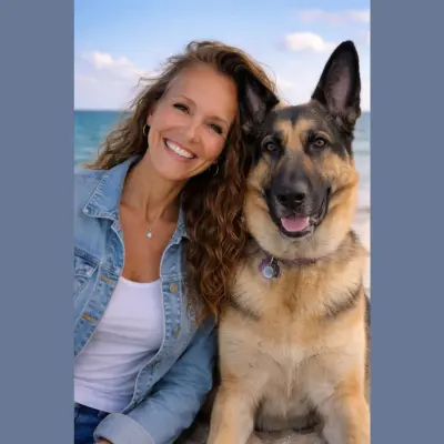 A woman with long, curly hair smiles while sitting next to a large German Shepherd on a sandy beach with the ocean and a clear blue sky in the background, embodying a joyful and serene seaside moment.