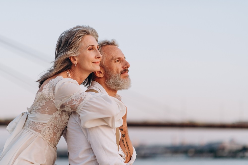 Grauhaariges Hochzeitspaar am Wasser im Abendlicht: Eine Person im Spitzenkleid umarmt die andere mit grauem Bart und tätowiertem Unterarm von hinten, beide blicken in die Ferne.