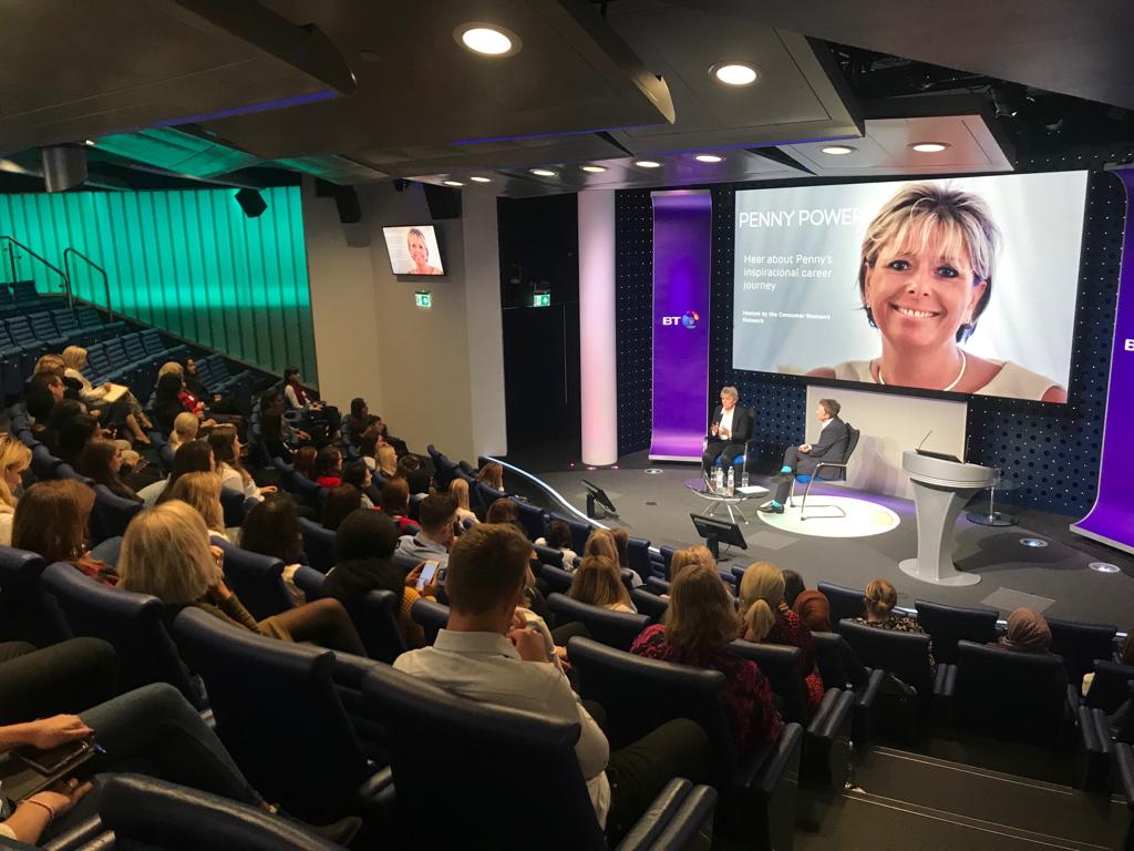 Cozy reading nook with sofa, chair, and plants inside a glass cabin with mountain forest views.