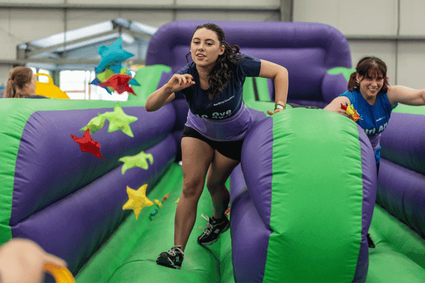 Outdoor team-building event with participants wearing matching blue shirts, taking part in inflatable obstacle courses and games, smiling and cheering, with the words 'ENTER A TEAM' in large bold white and green text across the image.