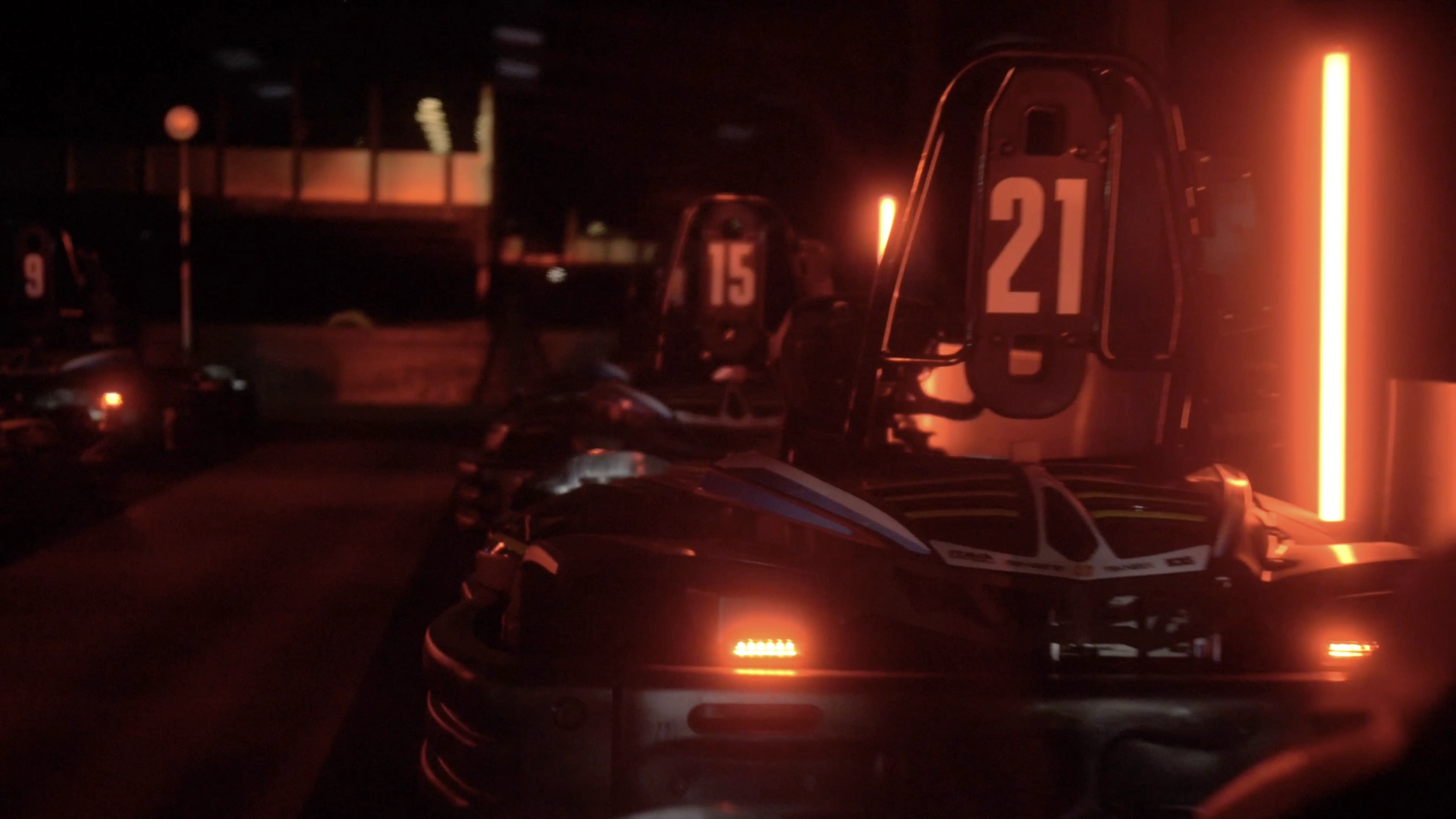 Dimly lit go-karts with illuminated numbers navigate a dark track, enhanced by glowing orange neon lights for a high-adrenaline, nocturnal racing ambiance.