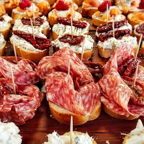 Un primer plano de varios aperitivos sobre pequeñas rebanadas de pan, cubiertos con carnes, quesos, tomates secos y guarniciones.